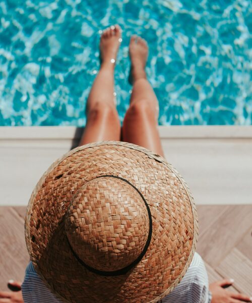 A woman wearing a straw hat with her feet in the water of a swimming pool seen from above